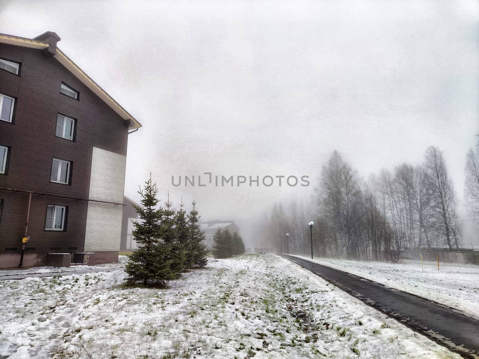 Snow blankets the ground as a dense fog envelops the pathway beside a residential building, creating a serene winter atmosphere.