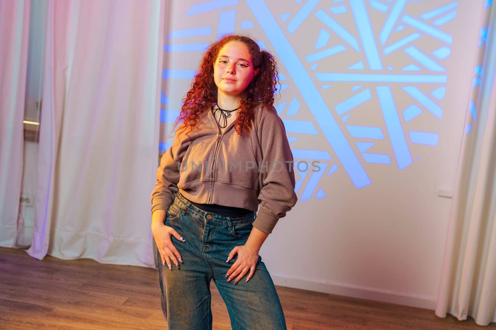 A young woman with red curly hair, wearing a brown hoodie and blue jeans, stands in front of a wall with a blue geometric light projection. She is smiling and looking at the camera.