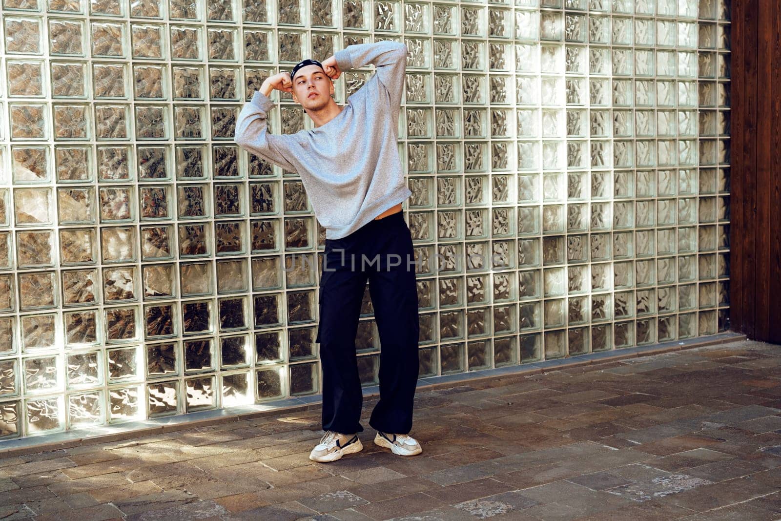 A young man, wearing a gray sweater and black pants, stands with his back to a glass block wall. The sunlight shines through the glass, creating a bright pattern on the ground.