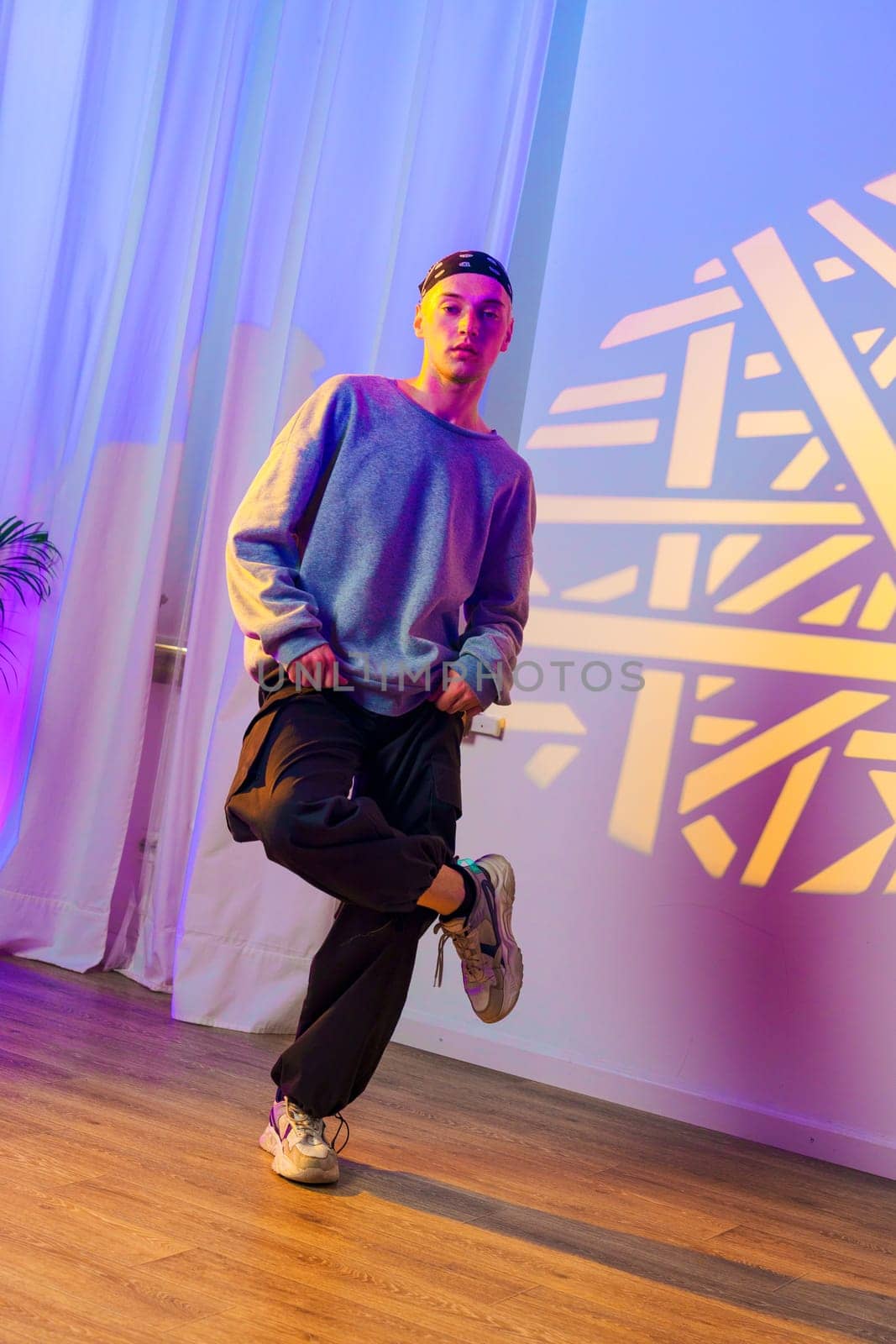 A young man, wearing a blue sweater and black pants, stands with one leg bent, his foot raised. He is in a dance studio, and a colorful light pattern is projected onto the wall behind him.