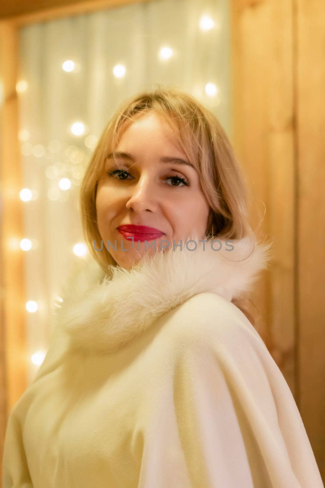 A woman with blonde hair and red lipstick smiles at the camera while wearing a white fur-trimmed coat. She stands in front of a wooden wall with string lights behind her.