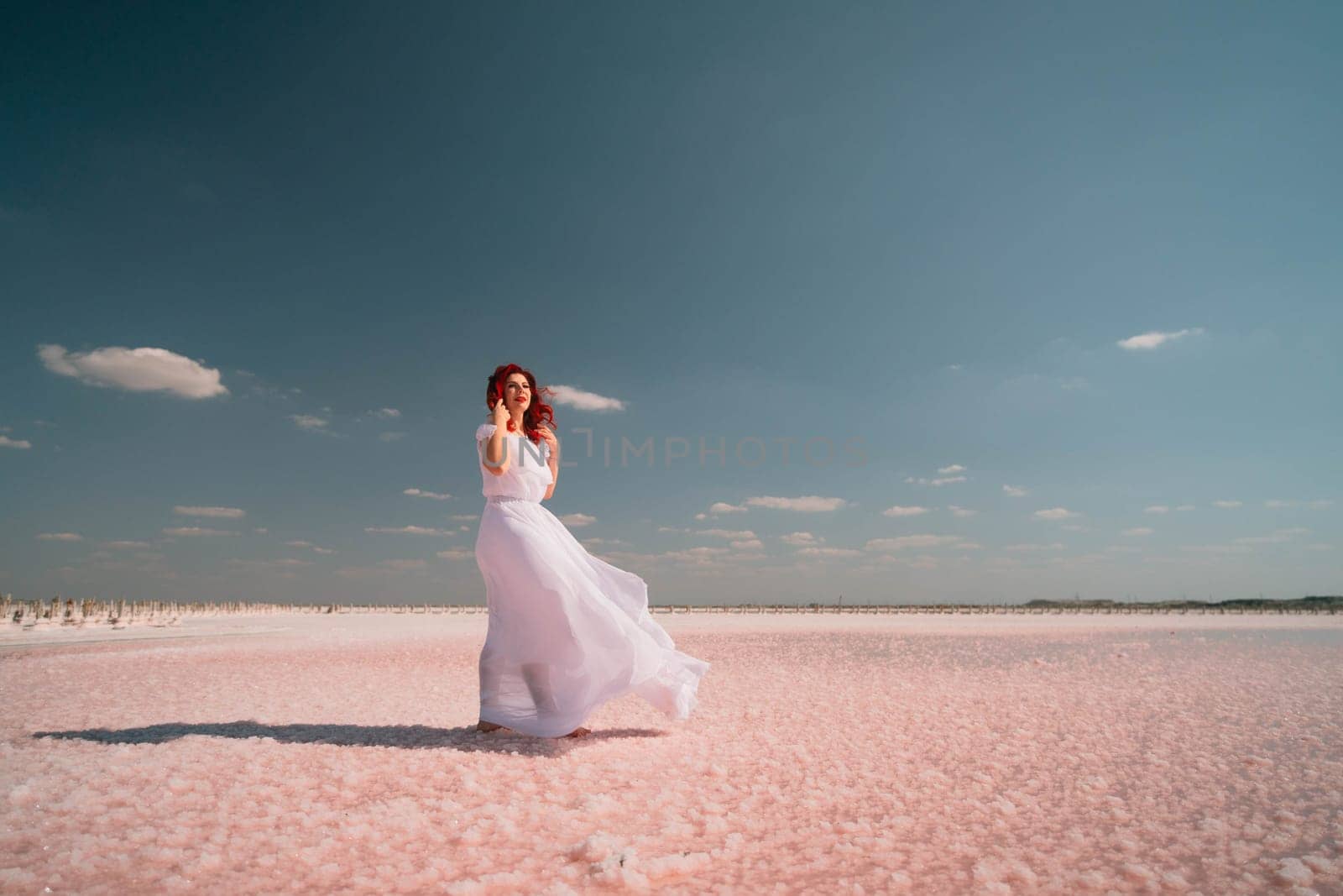 A woman in a white dress stands on a pink salt flat, her dress billowing in the wind. The sky is blue and cloudless.