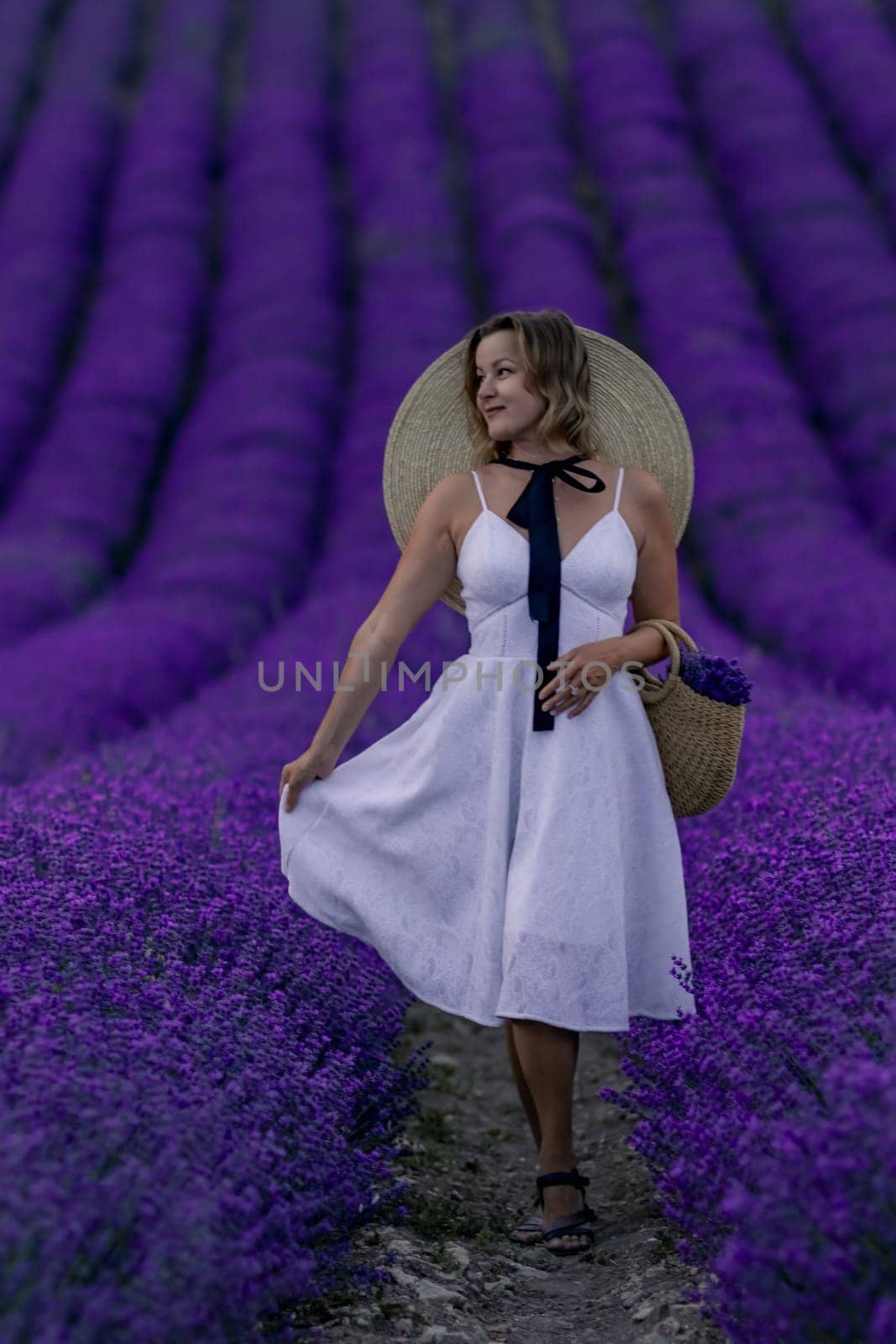 A woman in a white dress stands in a field of purple flowers. She is holding a basket and wearing a straw hat. The scene is serene and peaceful, with the woman enjoying the beauty of the flowers