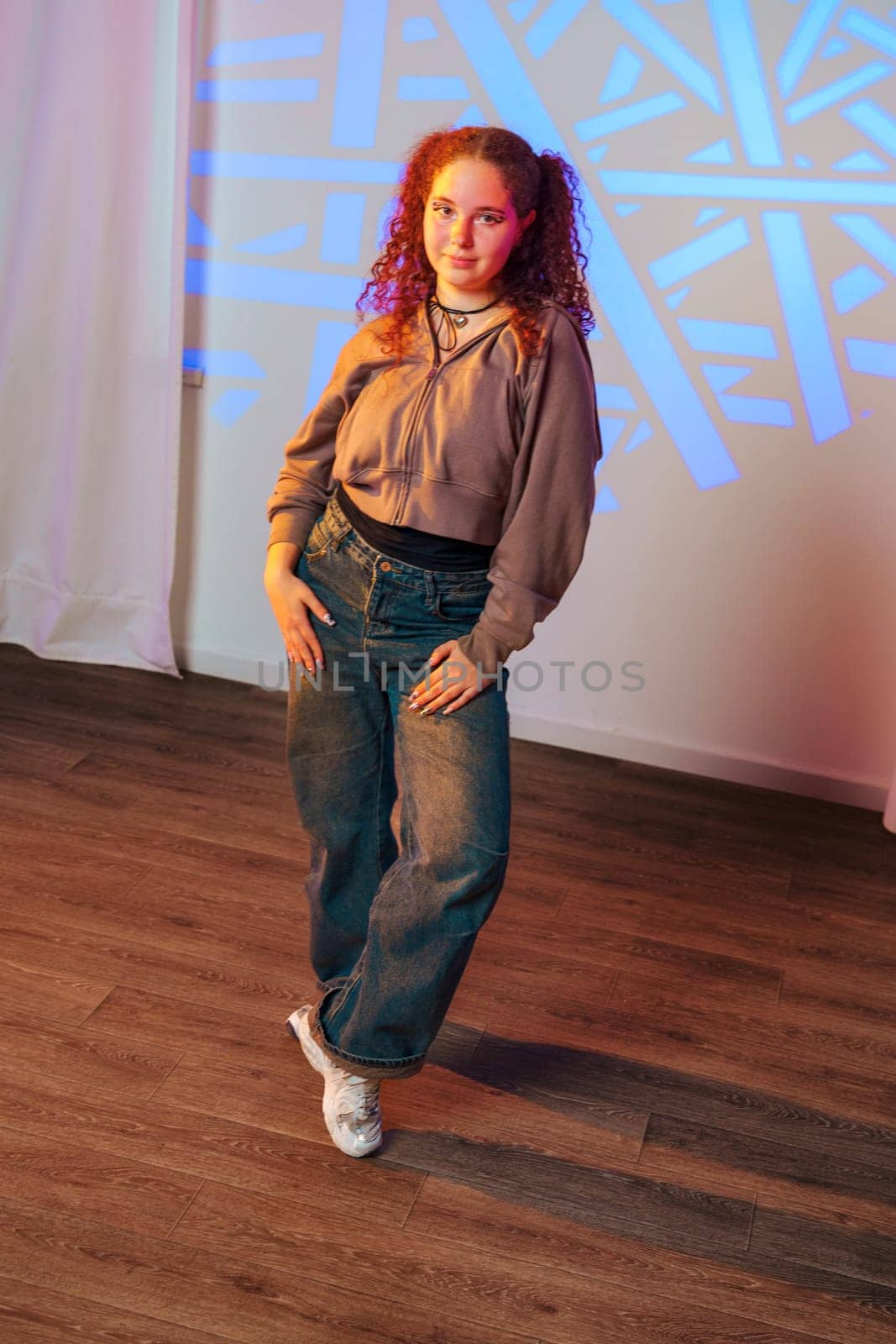 A young woman with curly red hair is standing in a dance studio. She is wearing a brown hoodie, jeans, and white sneakers. She is posing with one leg slightly raised and her hands on her hips.