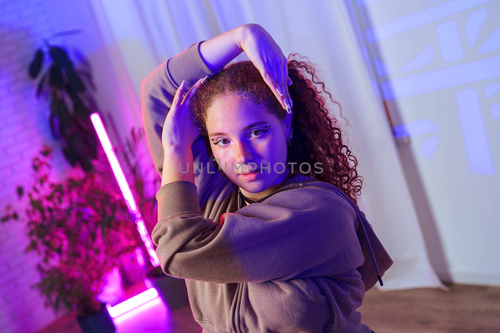 A young woman with curly hair and a grey hoodie is posing in a room with white walls and a pink neon light behind her. She is illuminated by a soft blue light.