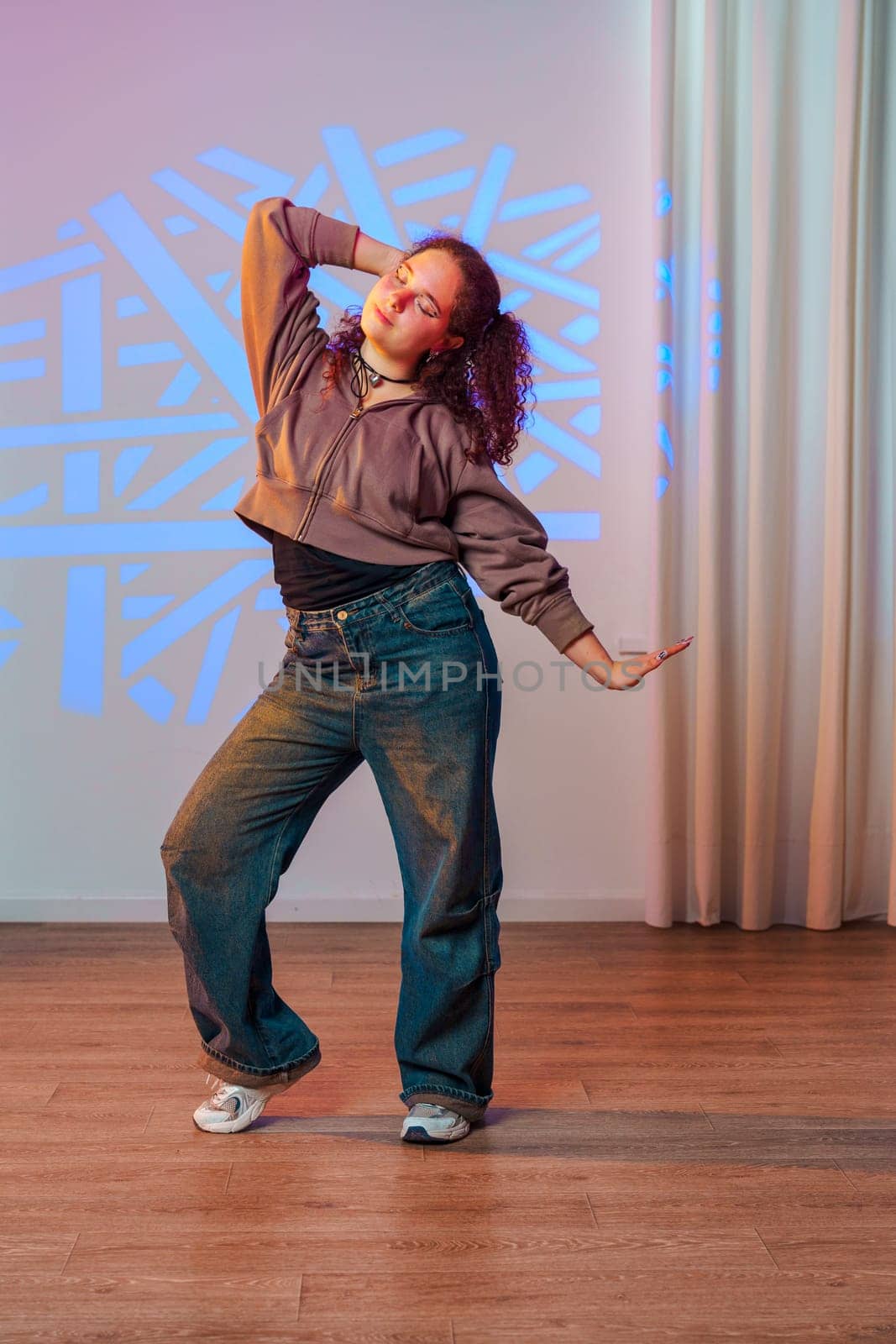 A young woman with curly hair dances in a studio. She is wearing a brown jacket, blue jeans, and white sneakers. The walls are white and there is a white curtain in the background.