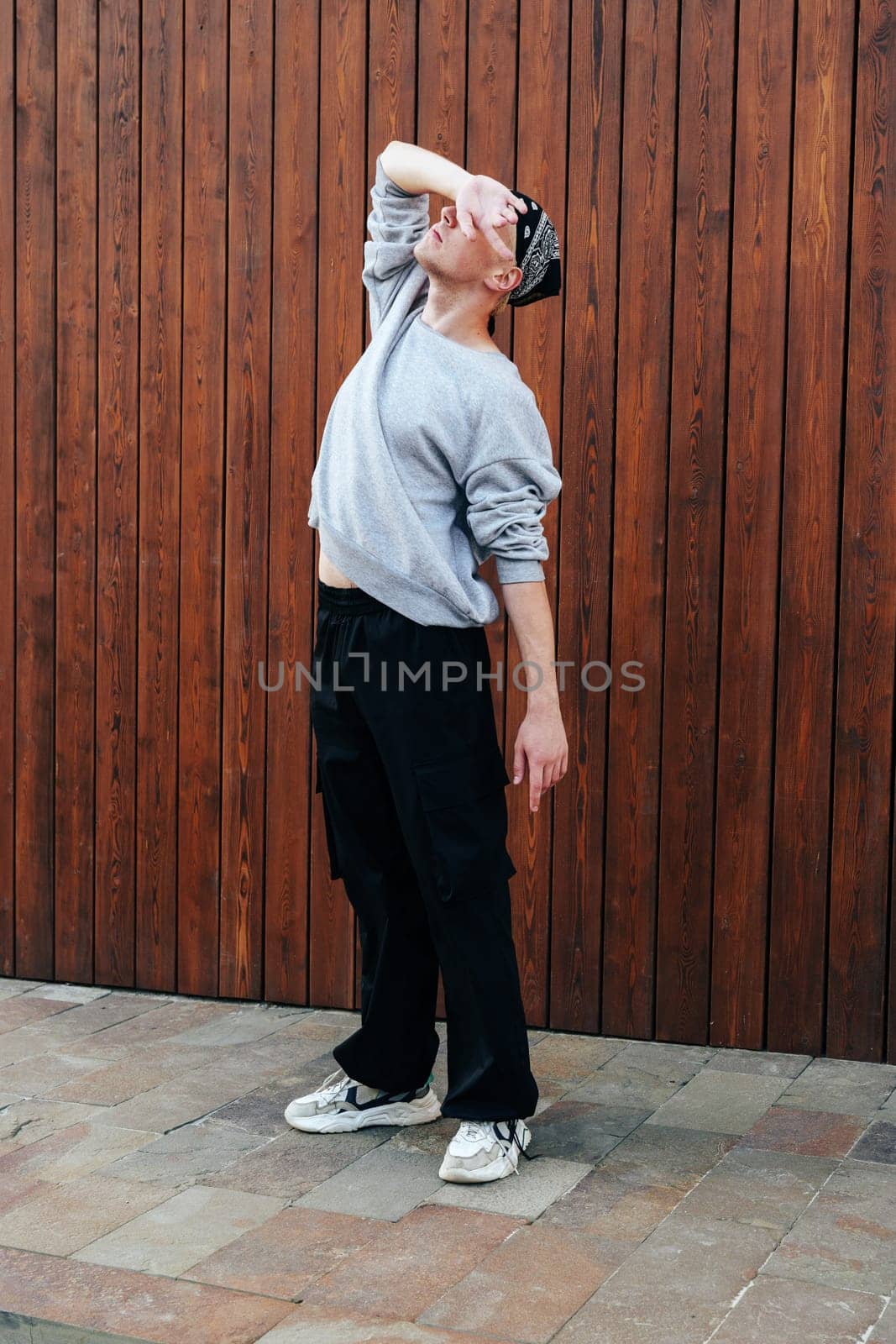 A person stands in front of a wooden wall with their hand over their eyes. They are wearing a grey sweatshirt, black pants, a bandana, and white sneakers.