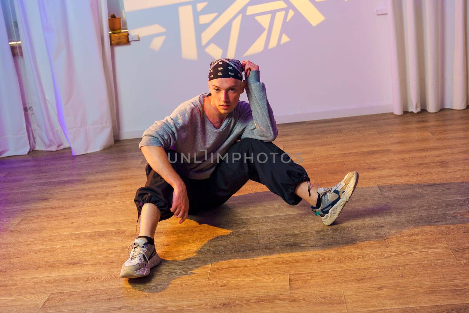 A young man in a bandana and sneakers sits on a wood floor in a dance studio. He has his right leg extended and his left leg bent at the knee. He is looking at the camera with a relaxed expression.