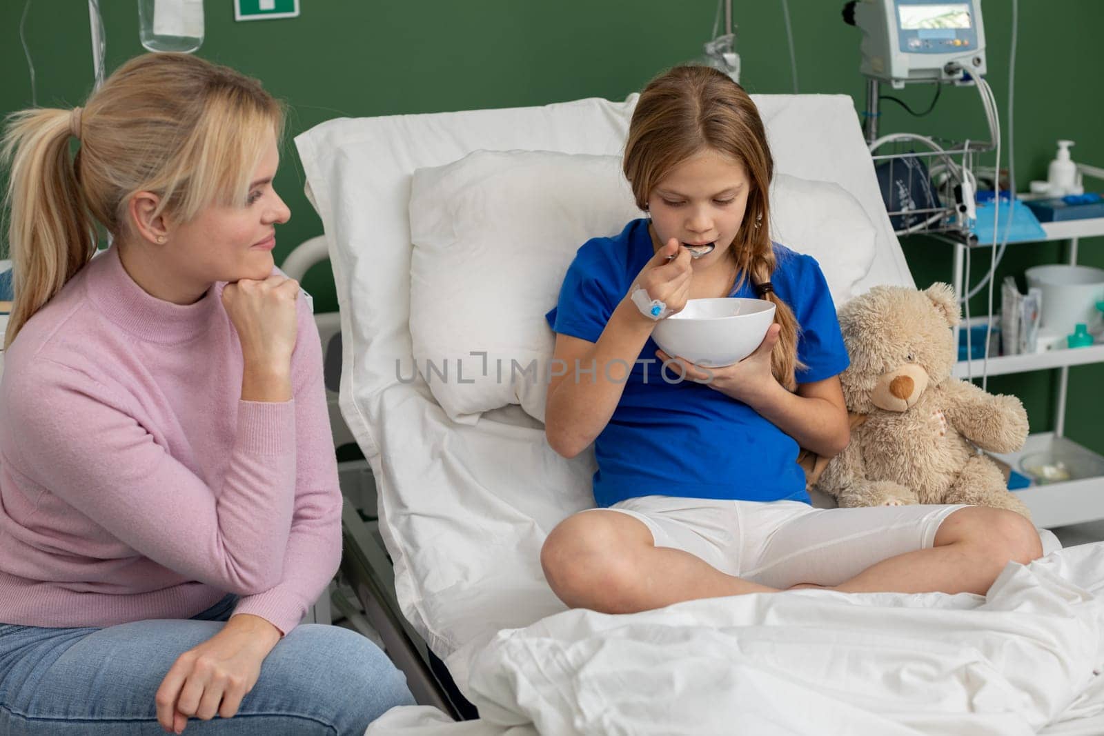 A young girl in a hospital bed is happily eating her meal while her supportive and caring relative looks on with love