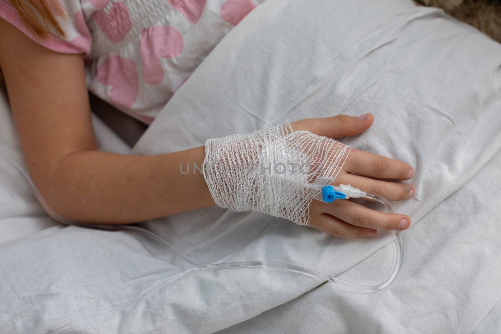 A small childs hand with an IV line and bandage gently rests on a soft pillow in a hospital setting