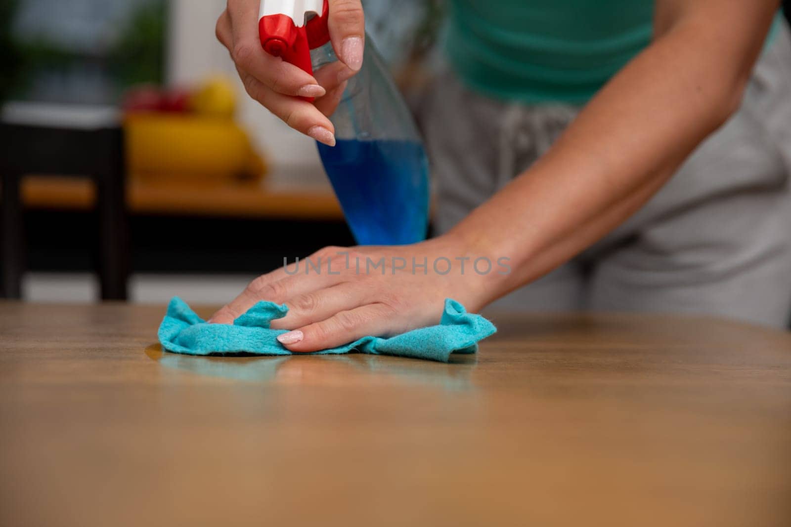 A woman is utilizing a bluecolored cleaning spray along with a cloth to carefully wipe and clean a wooden table