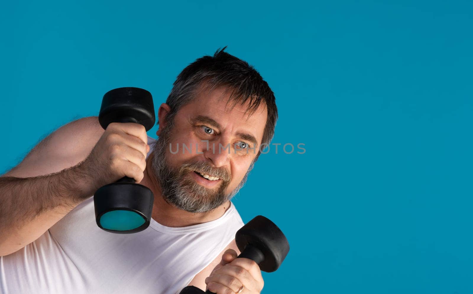 A middle-aged man with a beard lifts dumbbells in a vibrant blue studio, showcasing his determination and strength during an intense workout session aimed at improving fitness.