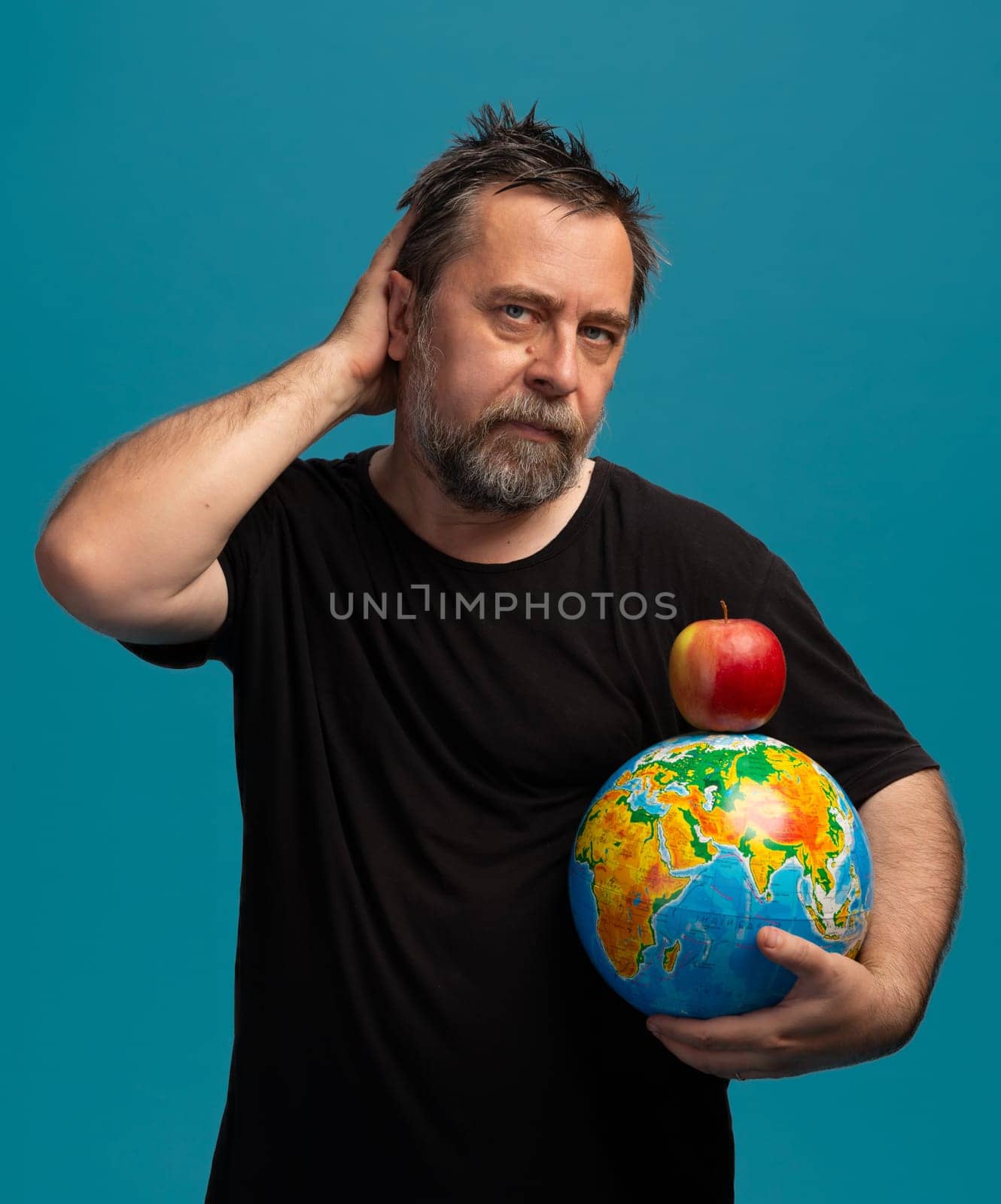 A man with a beard poses thoughtfully while cradling a globe in one arm and balancing a bright red apple on top. Set against a vibrant blue backdrop, he captures a moment of reflection.