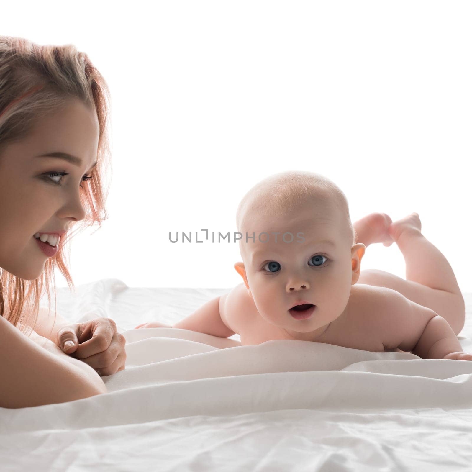 A mother smiles at her baby lying on a white surface, captured in a joyful interaction during a sunny day.