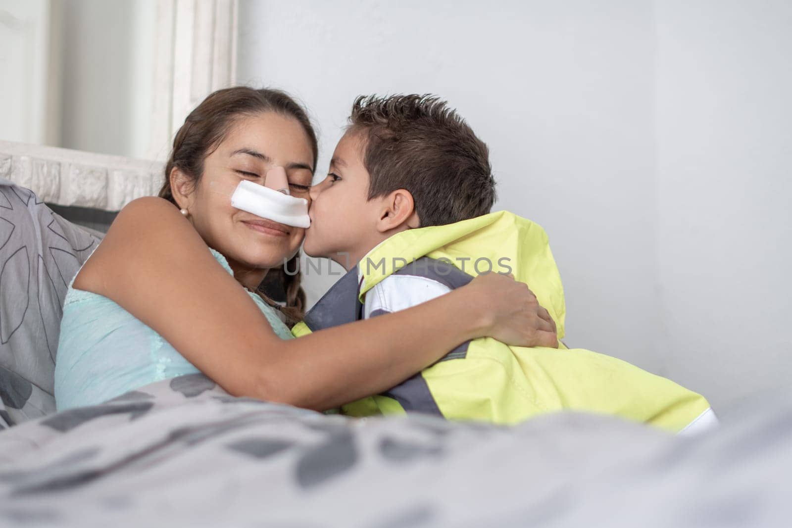 loving son hugging and kissing mother lying in bed after facial surgery, woman wearing gauze bandage on nose after rhinoplasty, support in difficult days, moments spent together by mother and child,