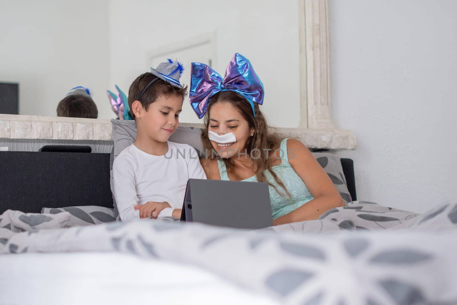 Smiling mother with bandage on nose lies in cozy bed and plays on tablet with son,mother has big blue bow on head and son has blue hat, carnival in bed to lift mood, relaxing,