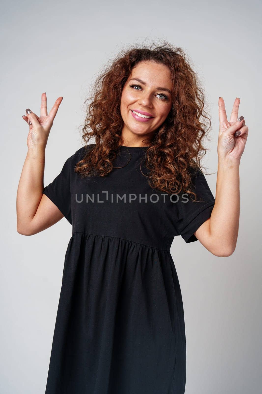 A young woman with curly hair poses cheerfully with peace signs in a neutral setting close up
