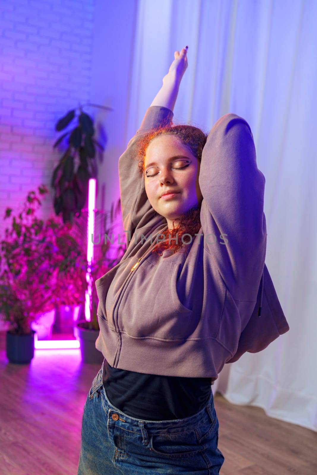 A young woman wearing a grey hoodie and blue jeans is stretching in a studio illuminated by colorful lights. She has red hair and is standing with her arms above her head.