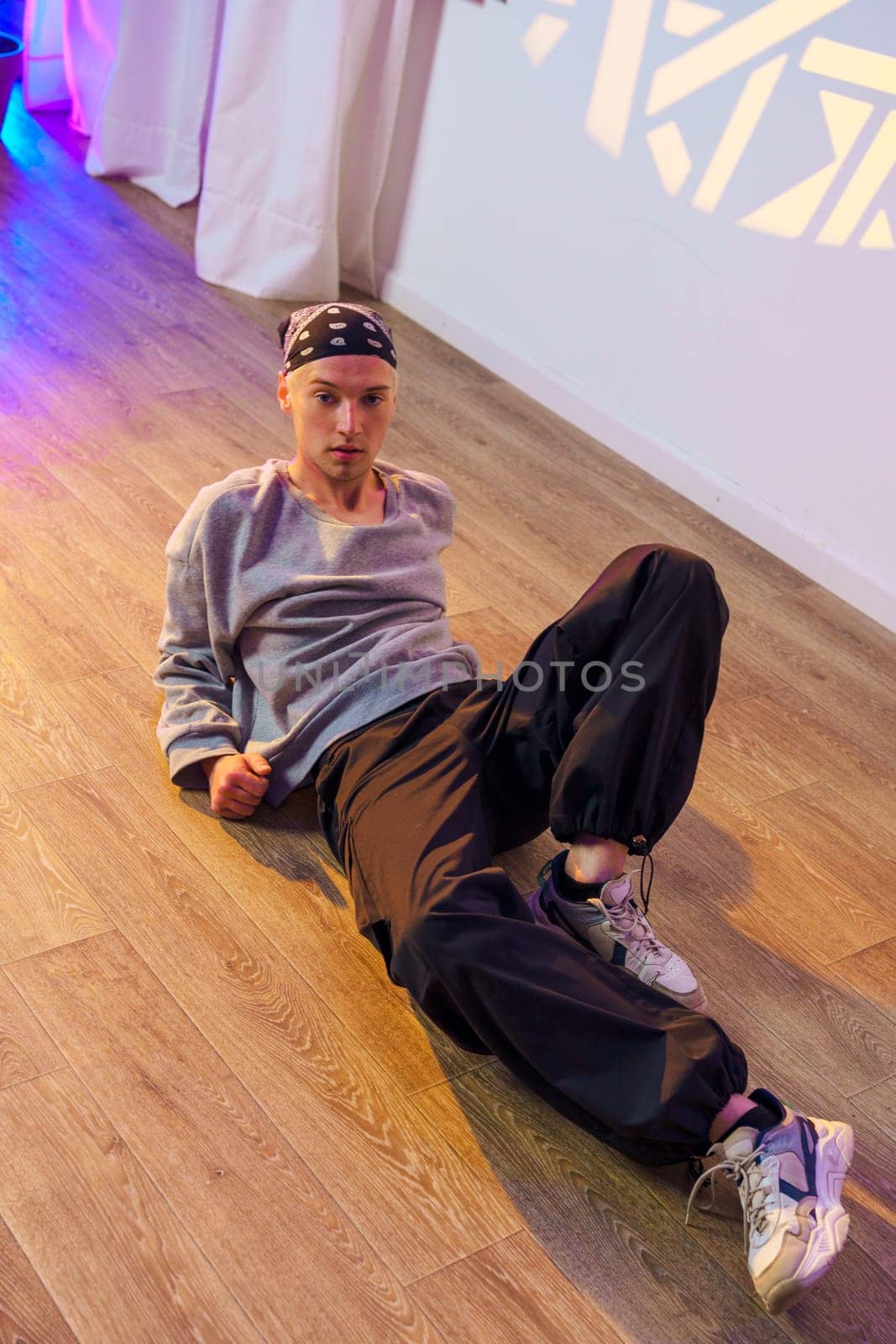 A young man, wearing a grey sweatshirt, black pants, and a bandana, is lying on a wood floor in a dance studio. The man's legs are outstretched, and he is looking directly at the camera.