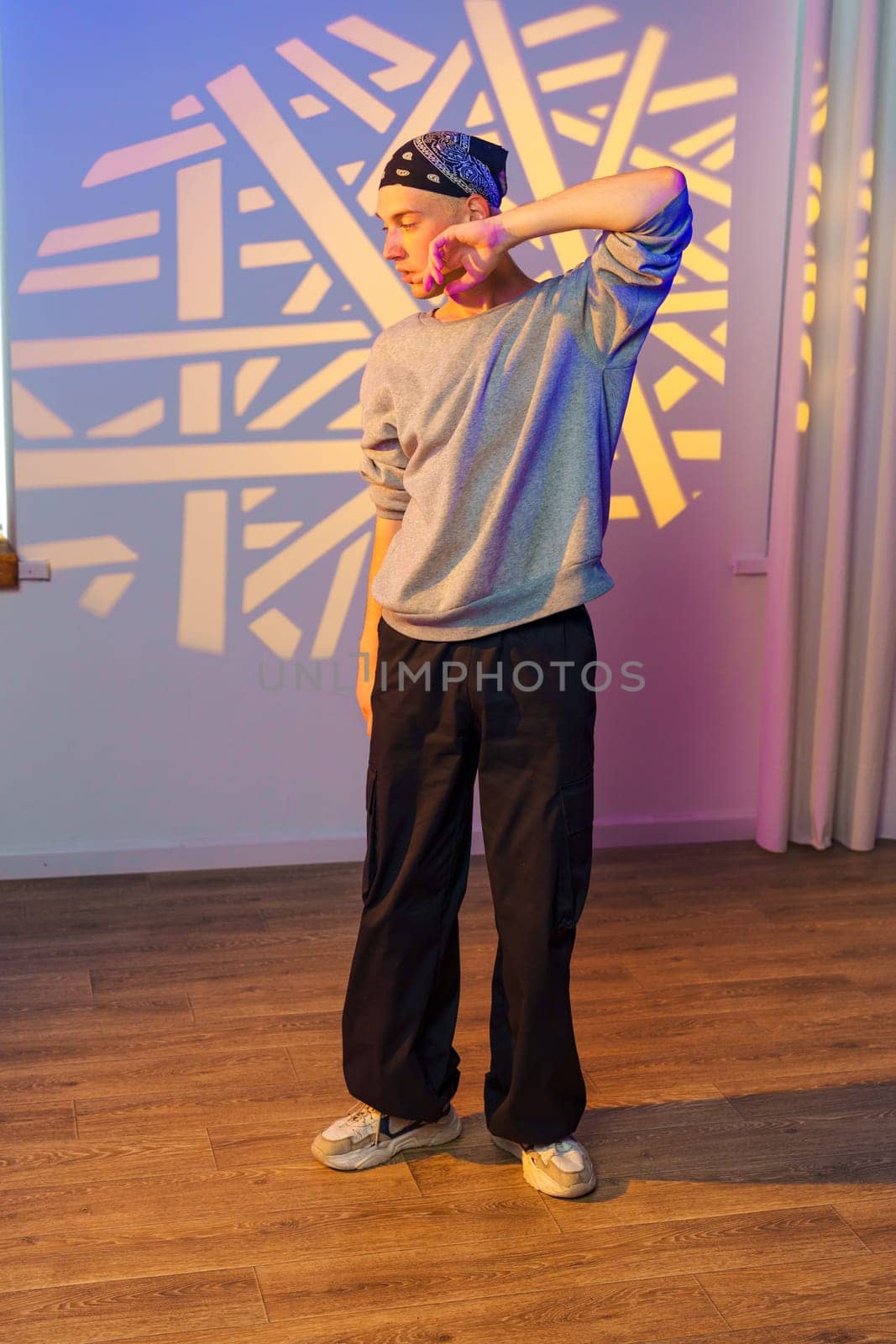 A young man in a grey sweatshirt, black pants, and white sneakers stands in a room. A geometric pattern of light is projected onto the wall behind him.