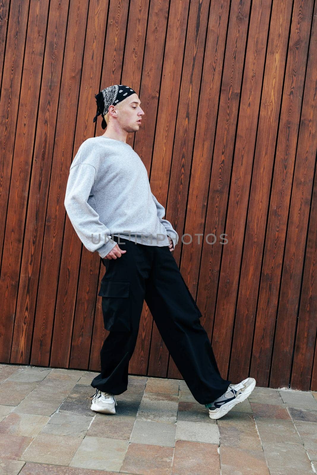 A young man wearing a grey sweatshirt, black pants, and a bandana walks past a wooden wall. The man's feet are slightly turned inward, and his hands are in his pockets.