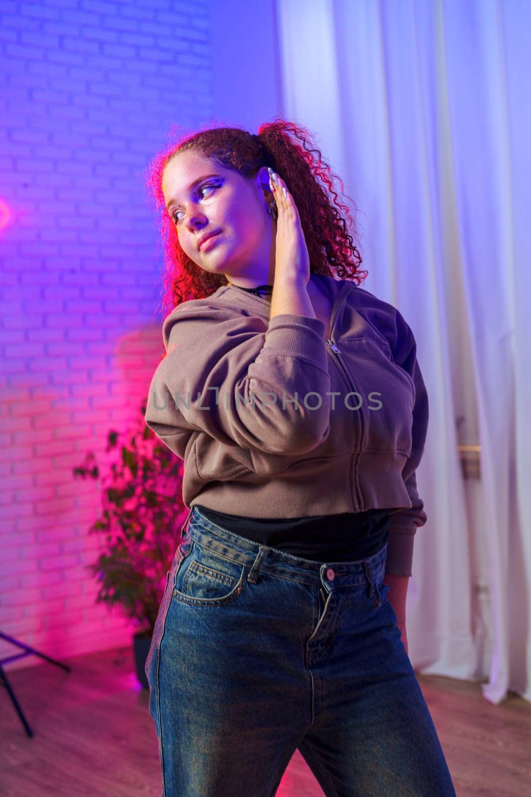 A young woman with long red curly hair is standing in a studio setting, wearing a brown jacket and jeans. She has one hand on her head and is looking off to the side.