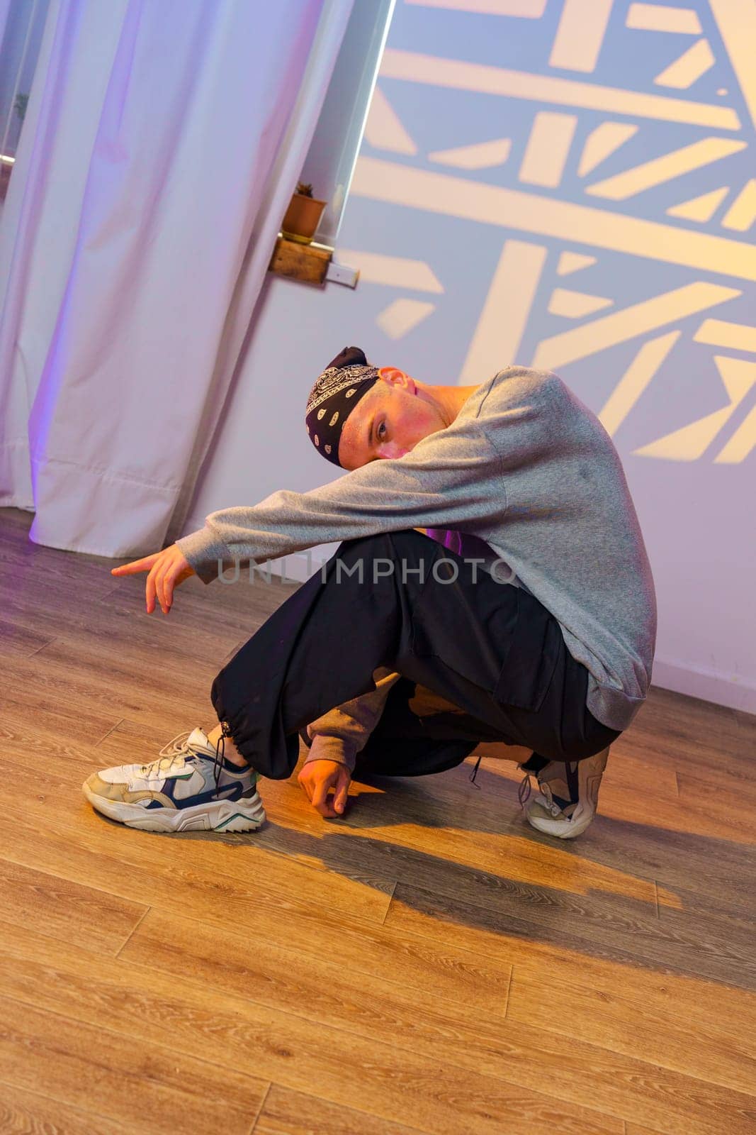 A young male dancer wearing a gray sweatshirt and black pants is in a low squat pose, looking towards the camera. The dancer's left hand is on the ground, while the right arm is outstretched.