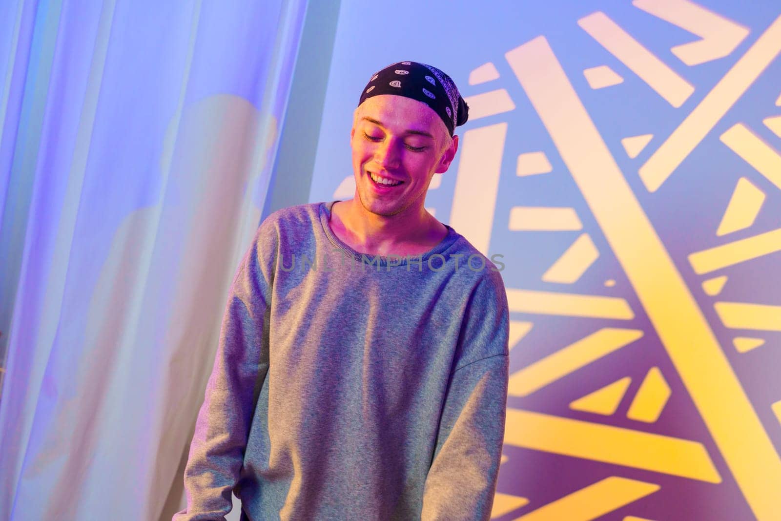 A young man with a bandana smiles as he stands in front of a blue and yellow patterned backdrop. The background is blurred, and the man is in focus. He wears a grey sweatshirt.