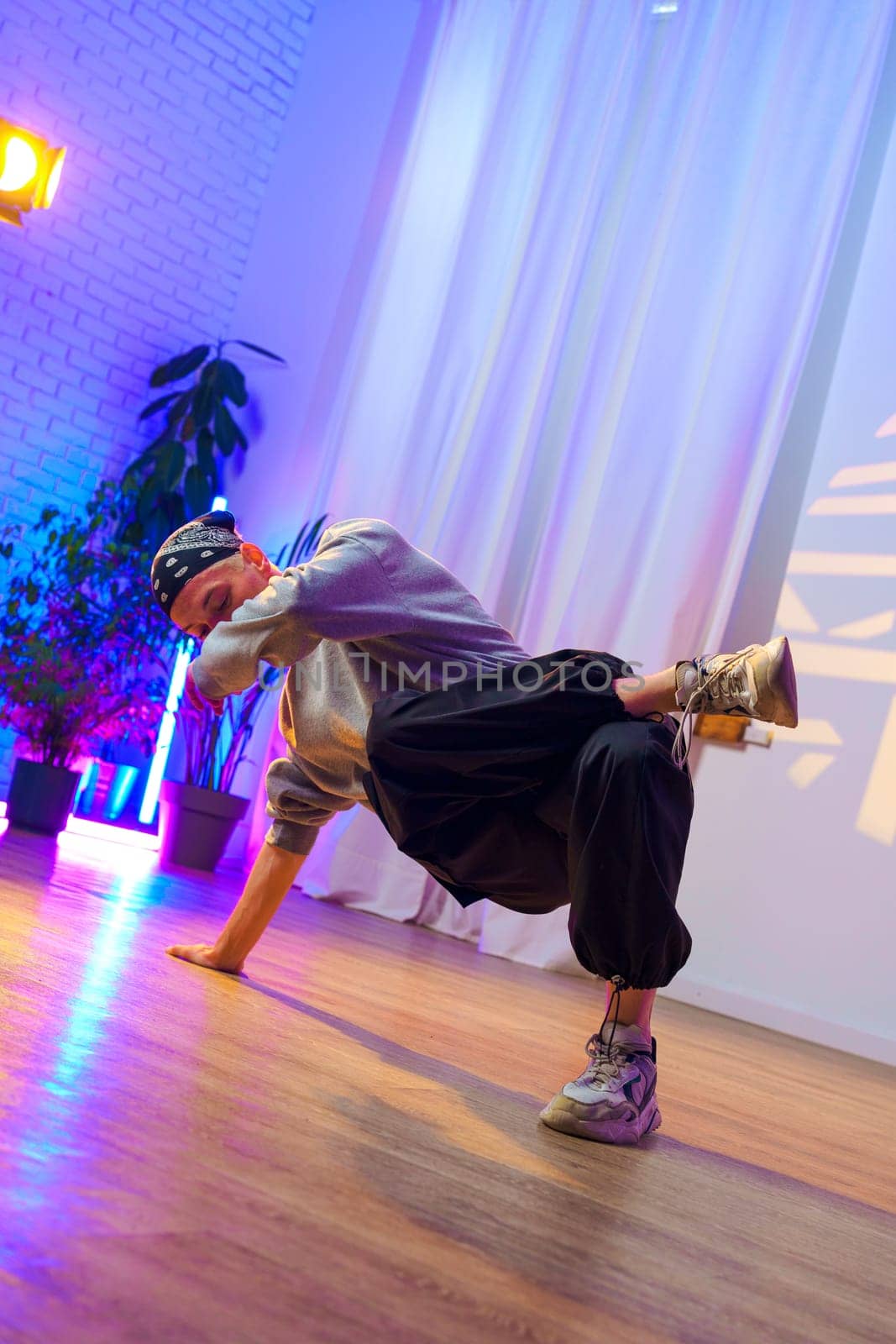A male dancer wearing a grey sweater, black pants, and a black bandana is performing a breakdancing move in a studio. The floor is wooden and there are plants in pots in the background.