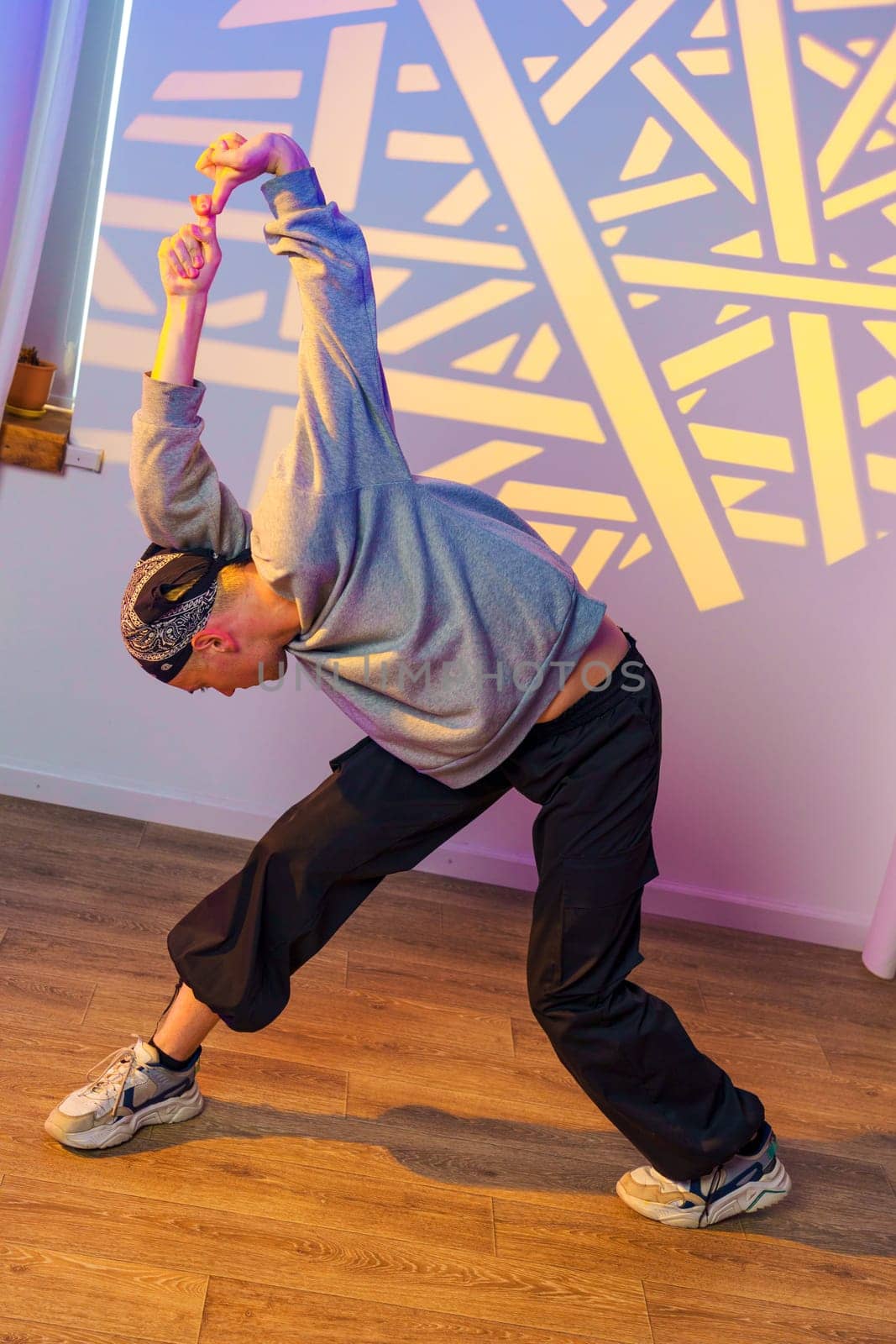 A dancer wearing a gray sweatshirt and black pants is stretching in a studio with a large geometric light projection on the wall. The dancer is bending over, reaching their arm towards the ceiling.