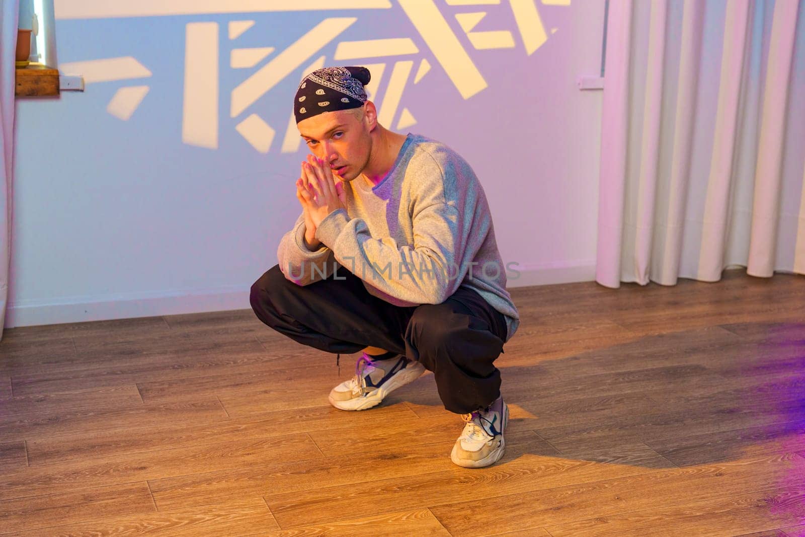 A young man is squatting on a wooden floor. He is wearing a grey sweatshirt and black pants. He has a bandana on his head and is looking at the camera.
