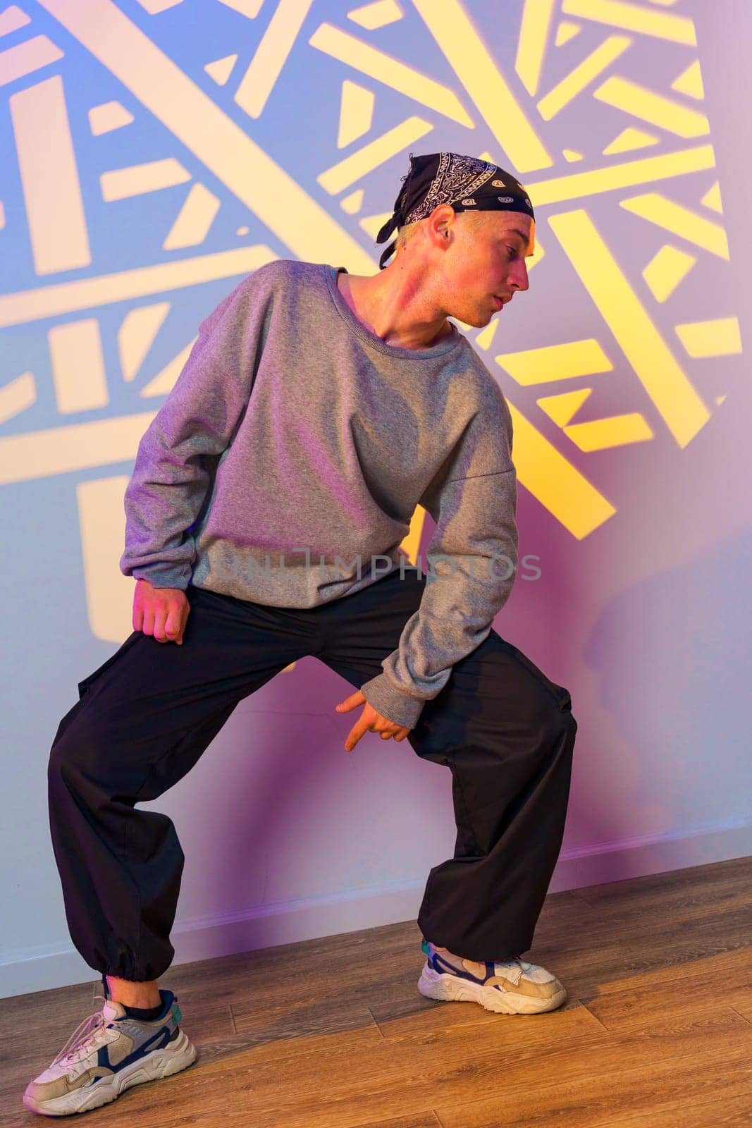 A young man wearing a grey sweater, black pants, and a bandana dances in a studio. The wall behind him is lit with a colorful light projection.