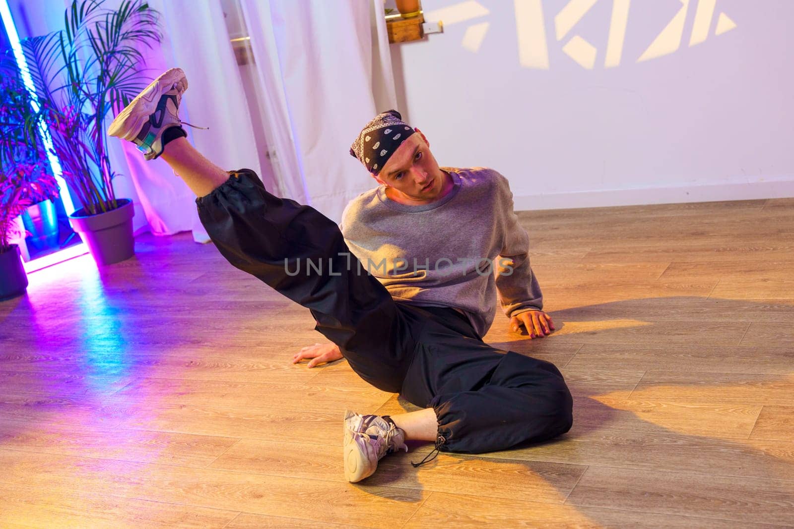 A dancer wearing black pants, a gray sweatshirt, and a bandana is in a dance studio. Their right leg is extended forward, and their left hand is on the floor.