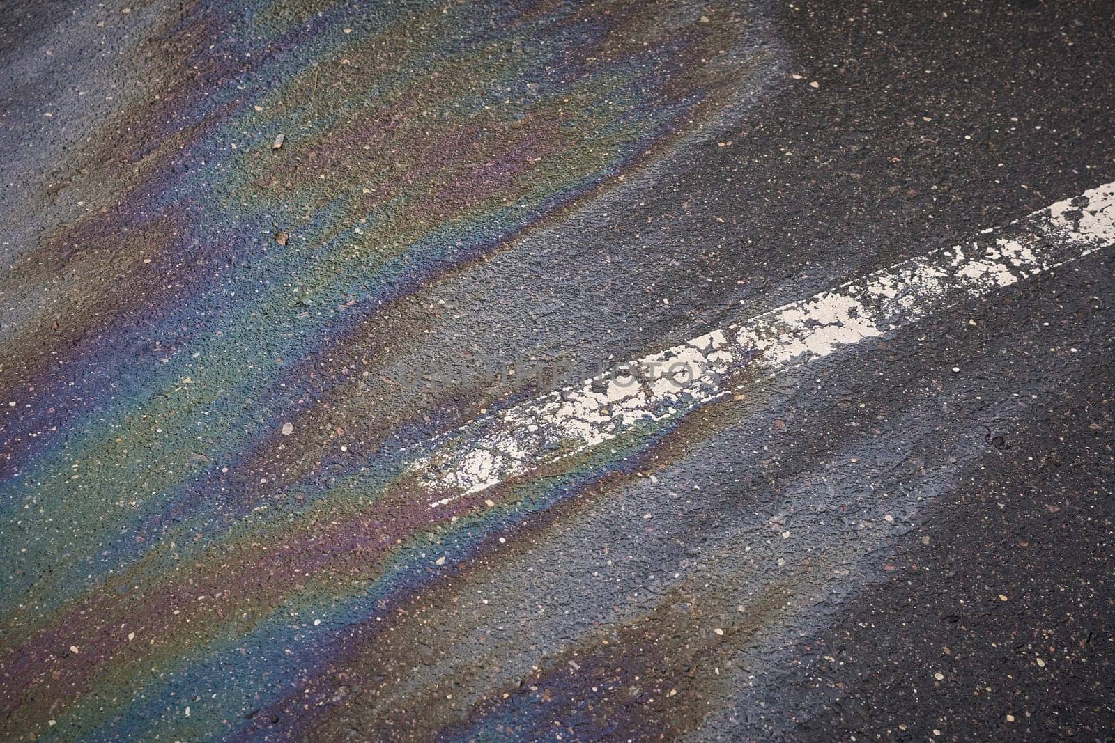 Colorful gasoline slick creating a shimmering rainbow effect on the dark asphalt, alongside a white road marking