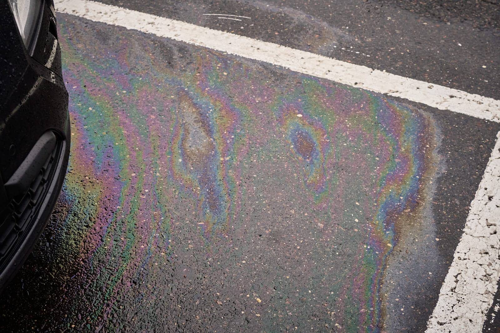 Multicolored gasoline stain generating a shimmering sheen on the wet asphalt near a vehicle, representing environmental pollution.