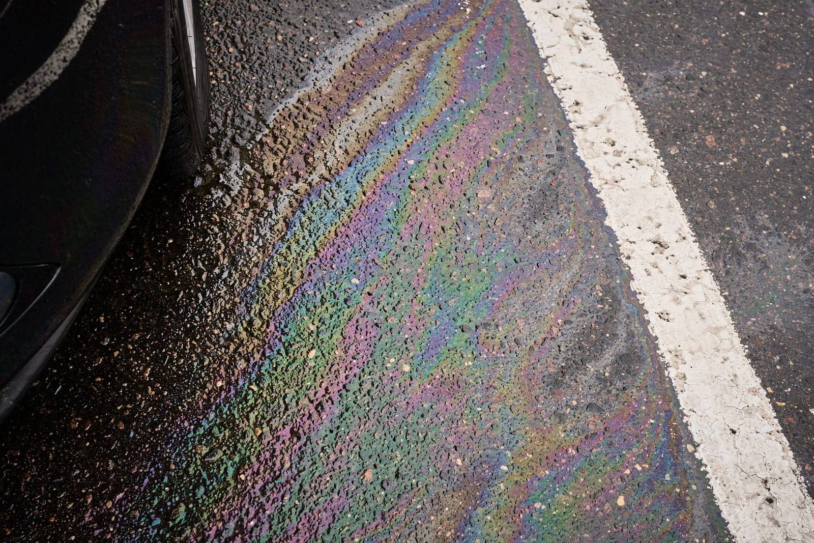 Colorful gasoline or oil spill creating a rainbow sheen on wet asphalt road near a car, illustrating environmental pollution