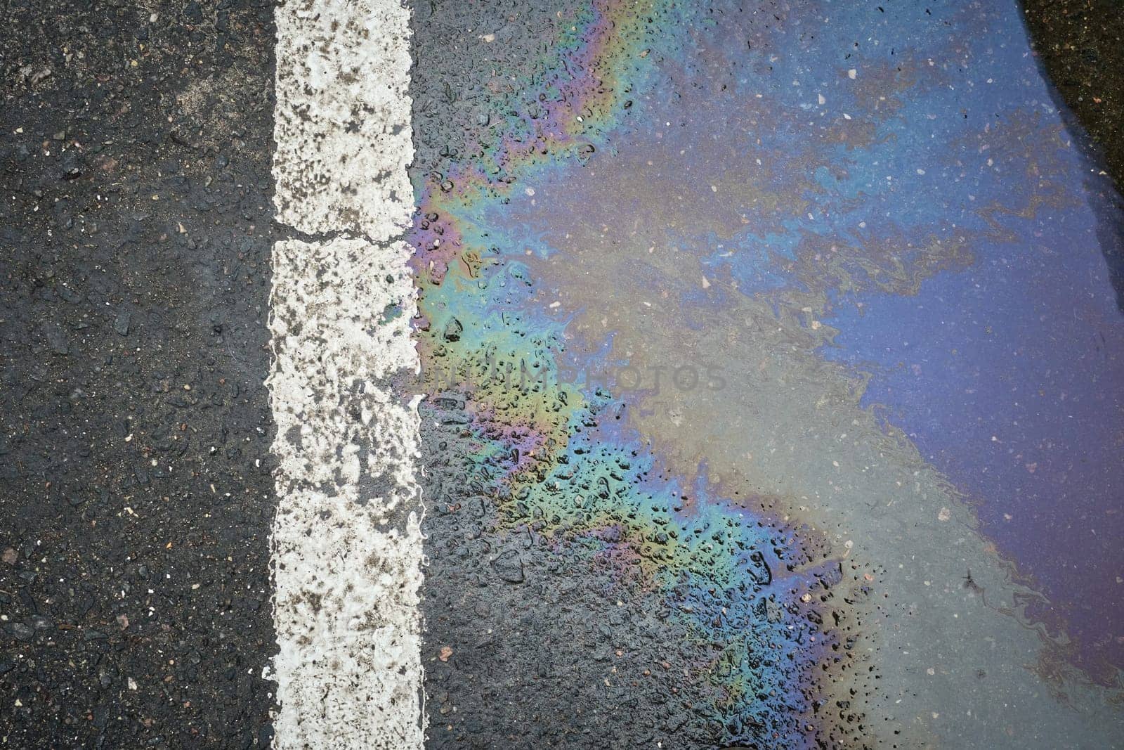Colorful gasoline or oil spill creating a rainbow sheen on wet asphalt road surface next to a white traffic line