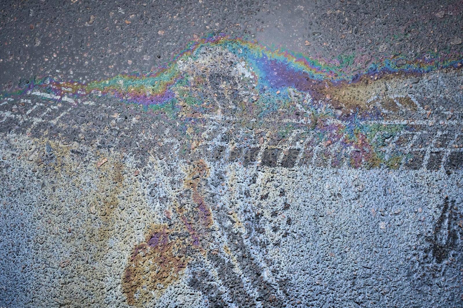 Spill of gasoline or oil with tire marks creates a rainbow sheen on the asphalt road surface.