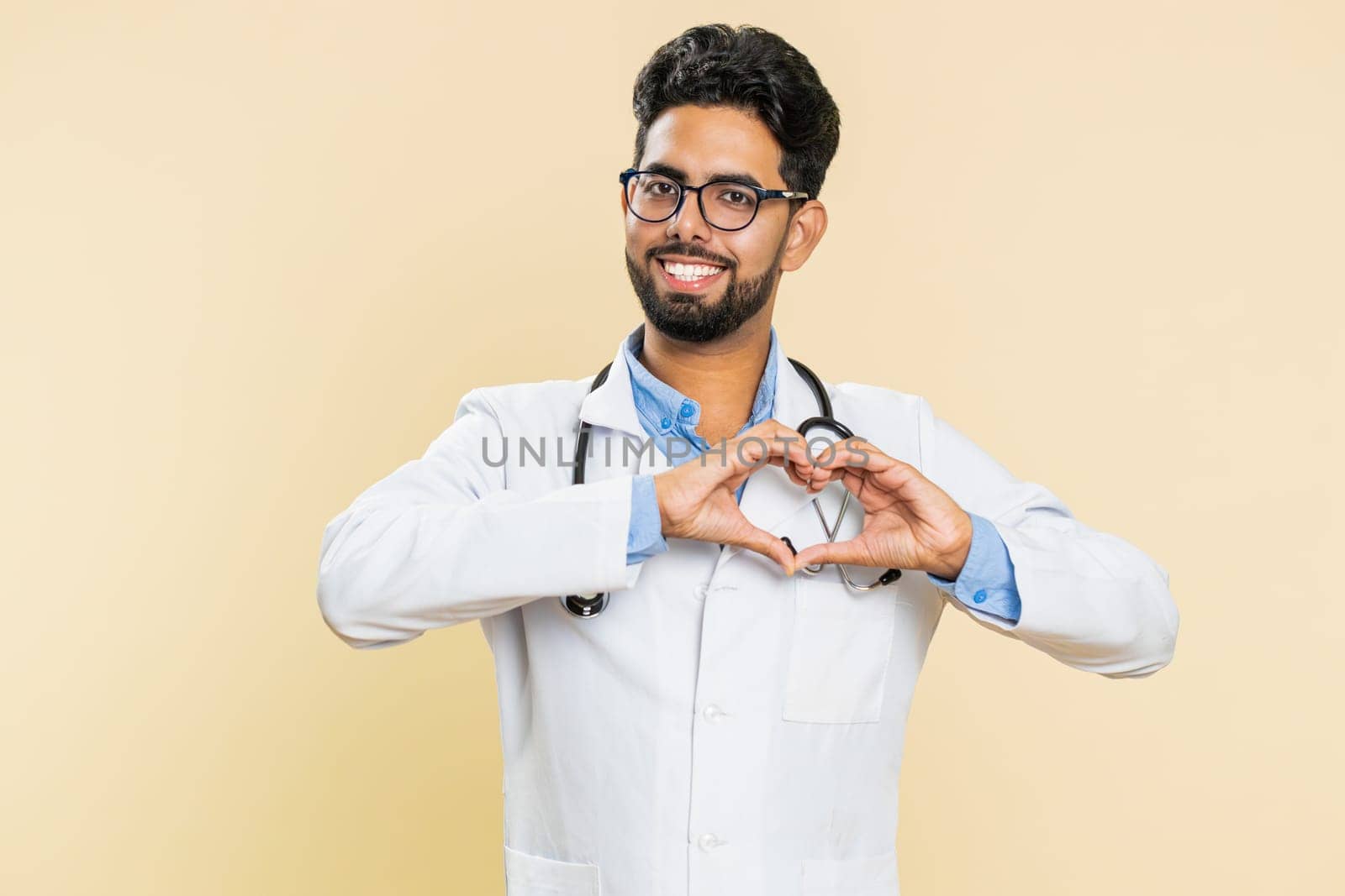 Indian doctor cardiologist man in love. Smiling attractive Arabian apothecary pharmacy guy makes heart gesture demonstrates love sign expresses good positive feelings isolated on beige background