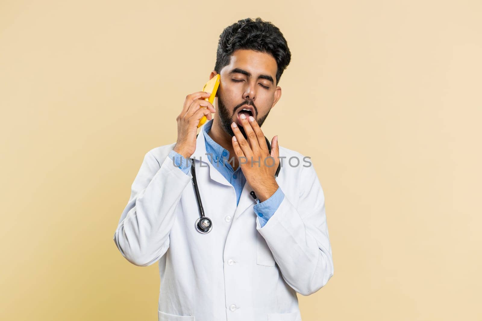 Disinterested Indian doctor cardiologist man having annoyed boring talk on smartphone. Tired sleepy Arabian apothecary pharmacy guy freelancer talking on mobile phone with friend on beige background
