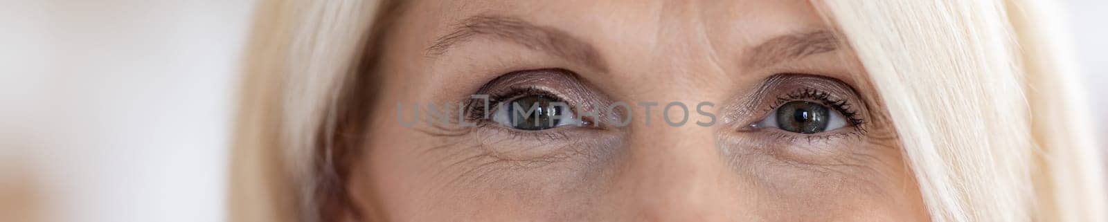 Eyes of attractive blonde senior woman in her 60s, blurred background, web-banner, panorama with copy space. Aesthetic medicine, plastic surgery, beauty treatment for elderly ladies