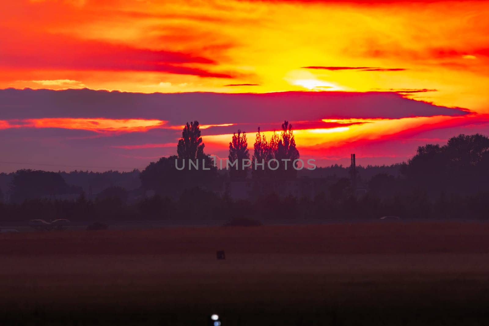 Beautiful cloudy red sunset with gigantic sun over big fields and trees seen by 600mm camera lens 