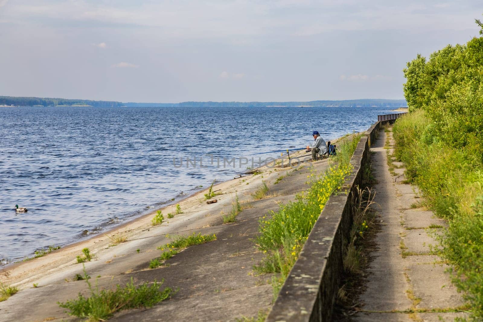 Old fisherman in hat and dark glasses do fishing at the coast of big lake at sunny morning by Wierzchu