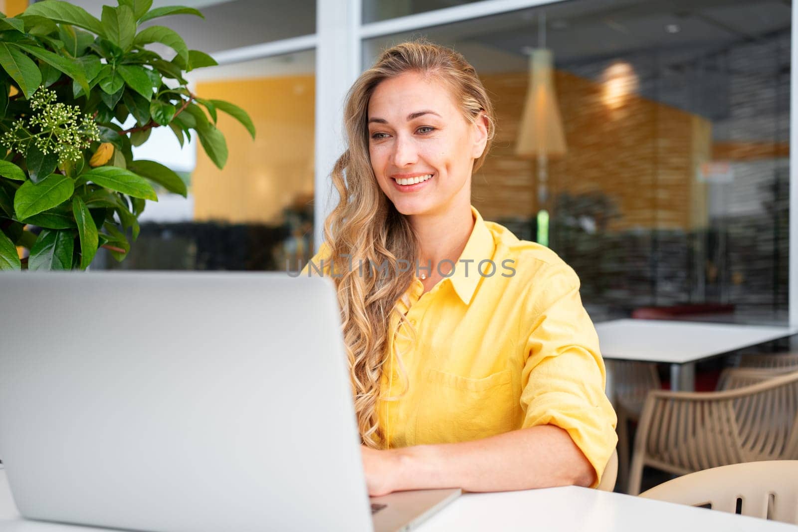 Freelance woman using laptop outdoor. Freelance worker sitting at cafe terrace remote working. Professional freelancer wearing yellow jacket sitting under green plant at cafe table with computer.