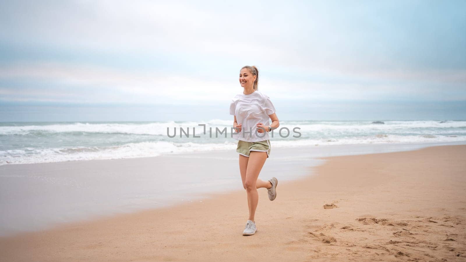 Woman jogging on beach. Sporty female jogging on sand beach, waves on background. Full body view woman in mini shorts and t-shirt who runs alongside ocean. Outdoor workout of trained fit girl.