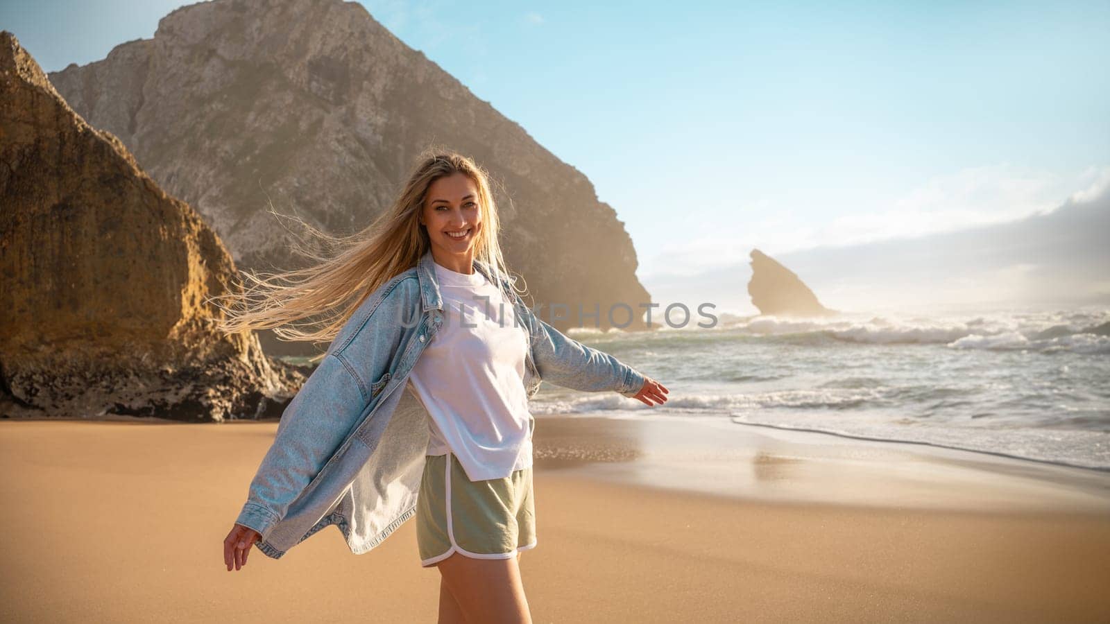 Woman enjoying being on beach with big rocks behind. Female with long blond hair is happy and smile while dancing on sand near ocean. Girl in short and denim jacket smiles to the camera on sea coast