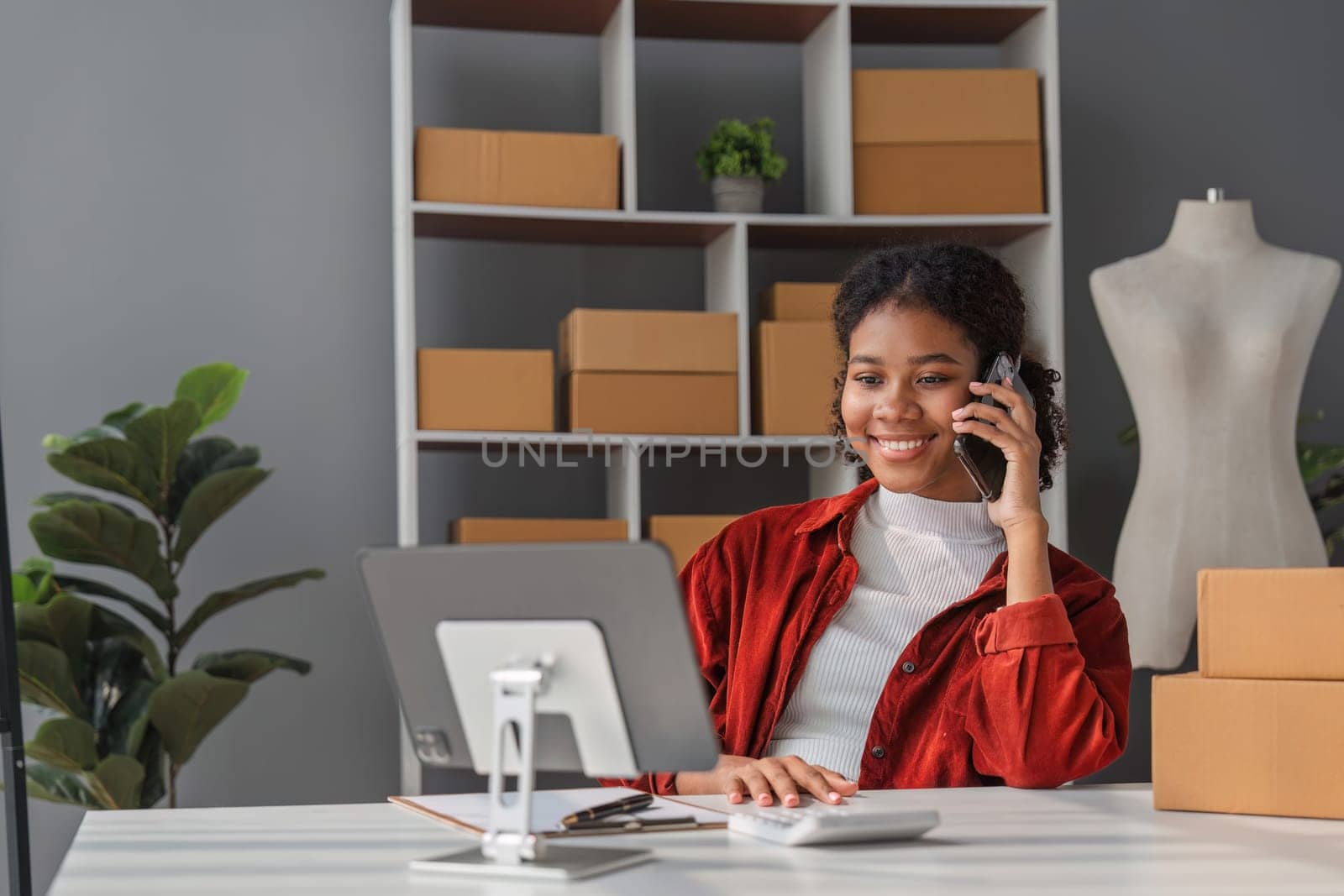 A small SME entrepreneur communicates business via a tablet and talks on the phone in an office with boxes of goods ready for delivery..