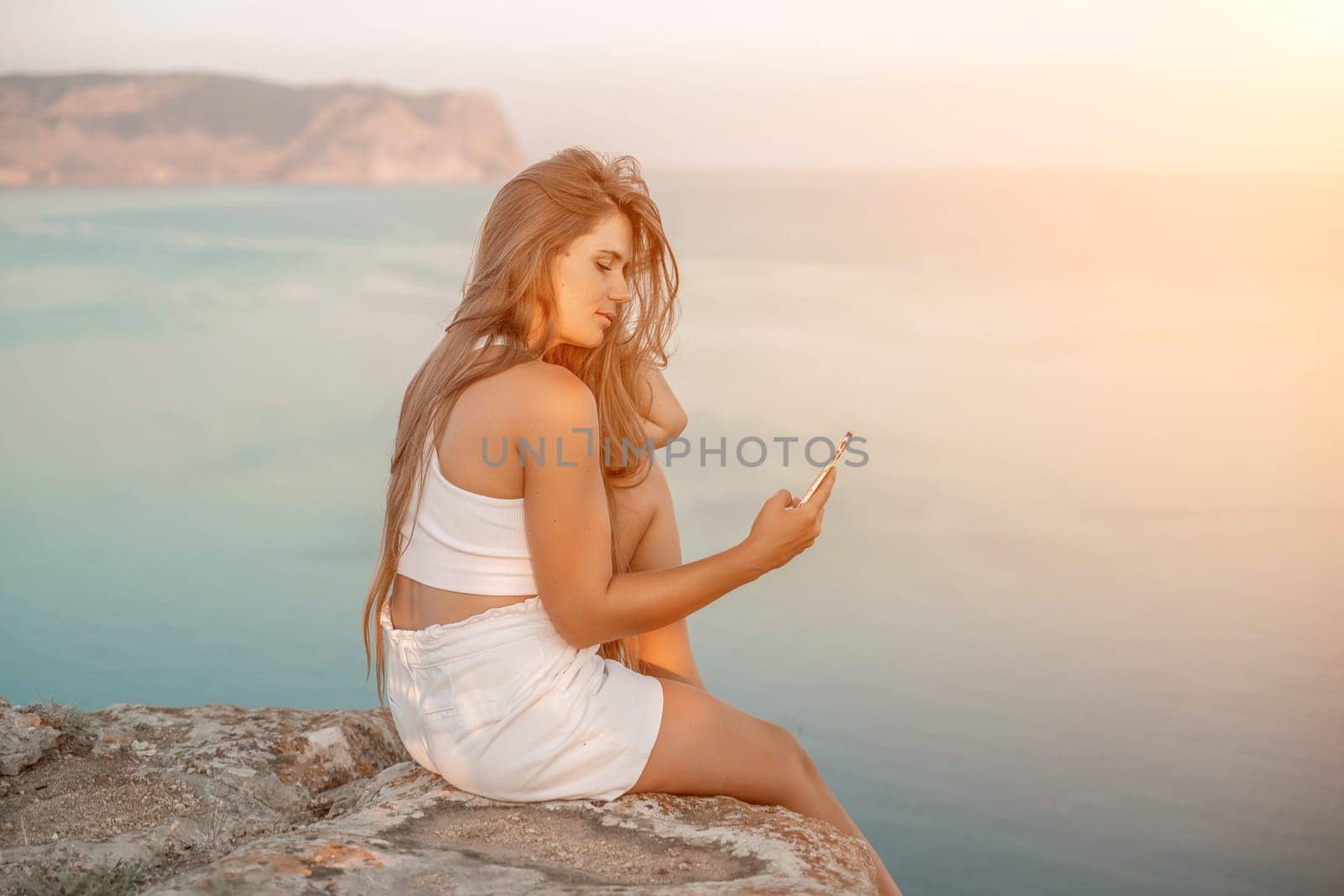 Happy woman in white shorts and T-shirt, with long hair, talking on the phone while enjoying the scenic view of the sea in the background