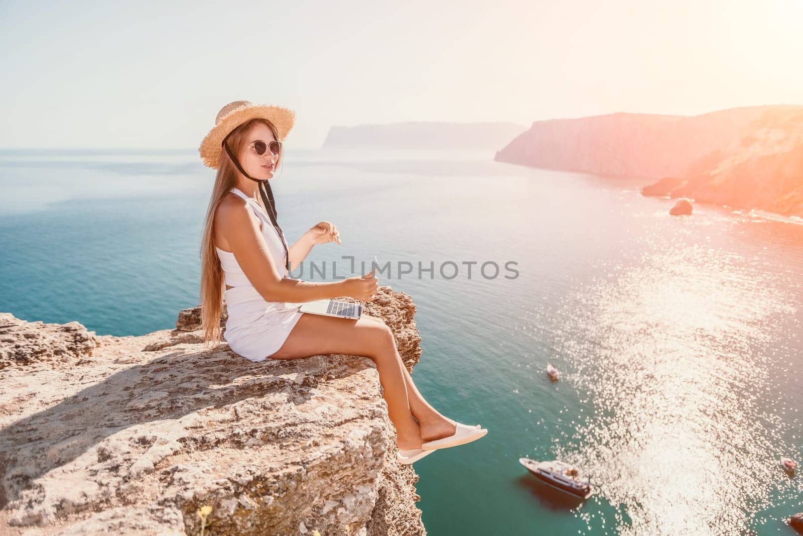 Woman laptop sea. Working remotely on seashore. Happy successful woman female freelancer in straw hat working on laptop by the sea at sunset. Freelance, remote work on vacation by panophotograph