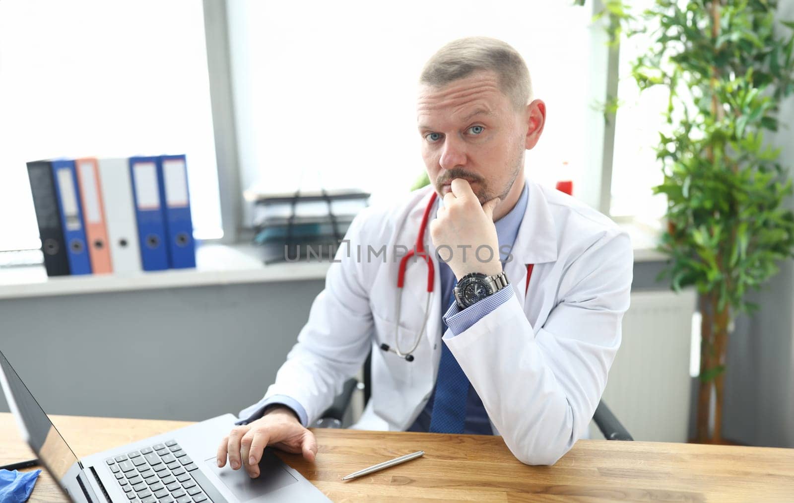 Portrait of pensive doctor at workplace with laptop in clinic by kuprevich